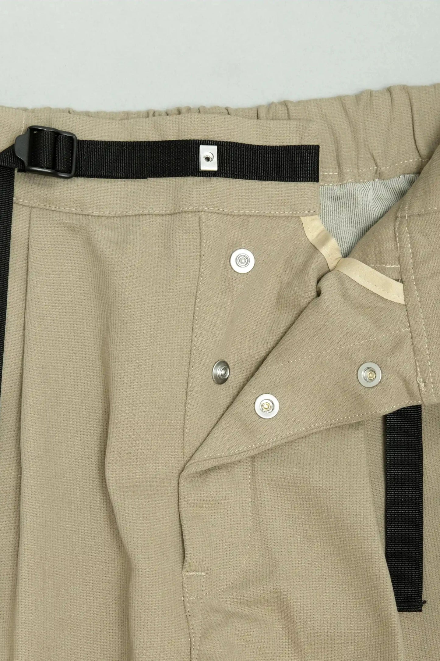 Close-up of beige relaxed fit pleated pants made from durable fabric featuring a black adjustable belt and snap button fasteners, highlighting utility-inspired craftsmanship.