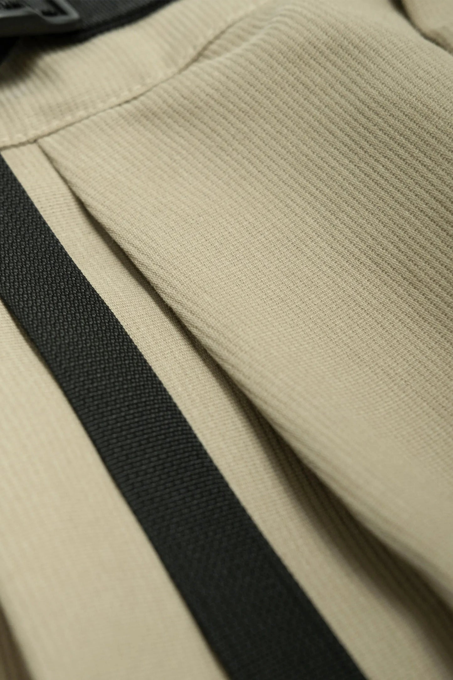 Close-up of beige textured fabric with black strap, showcasing detailed stitching and craftsmanship in a relaxed fit military-inspired pant design.
