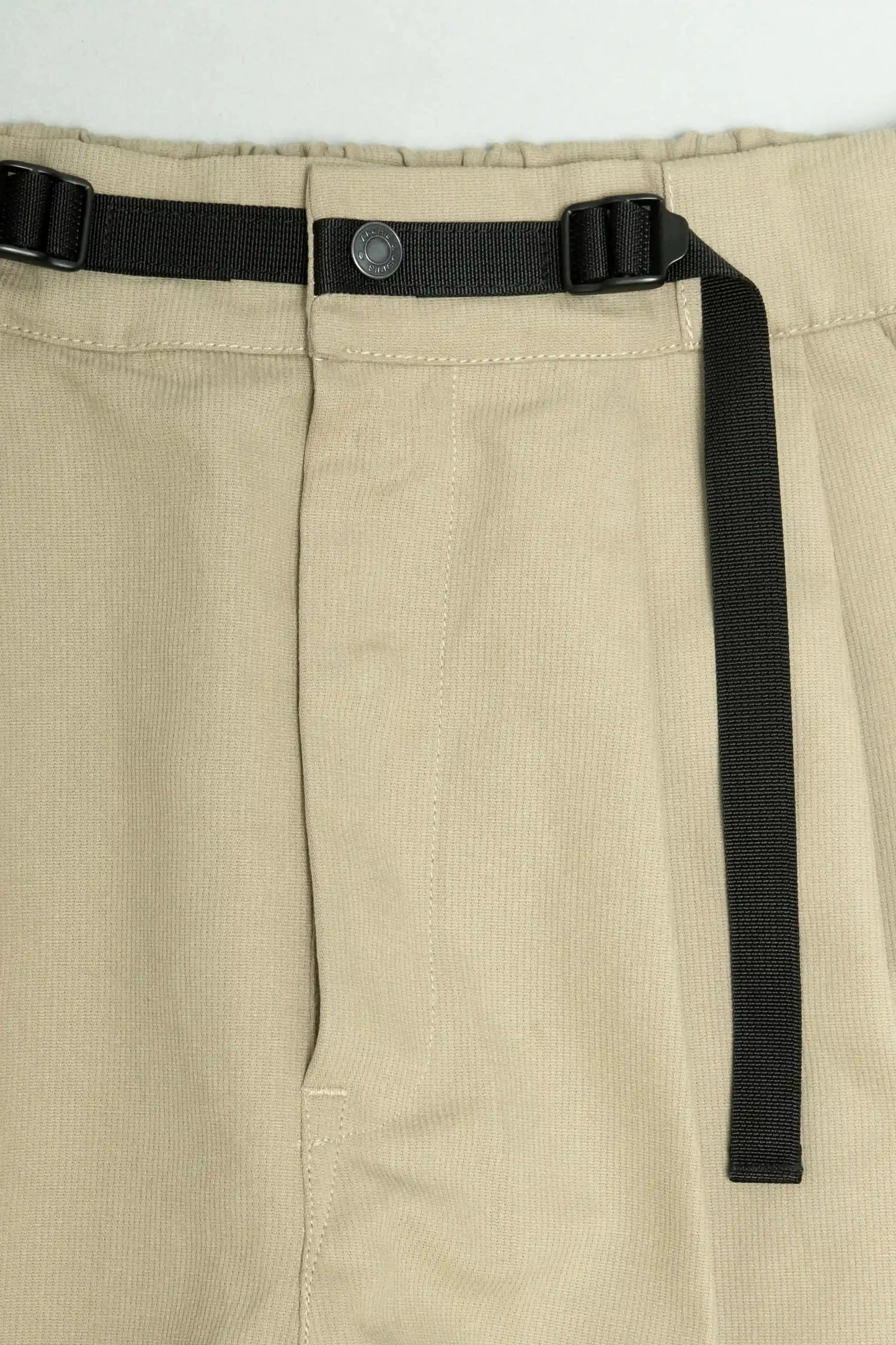 Close-up of beige pleated pants featuring a black adjustable utility-style belt with buckle and snap button closure, showcasing relaxed fit military-inspired menswear details.