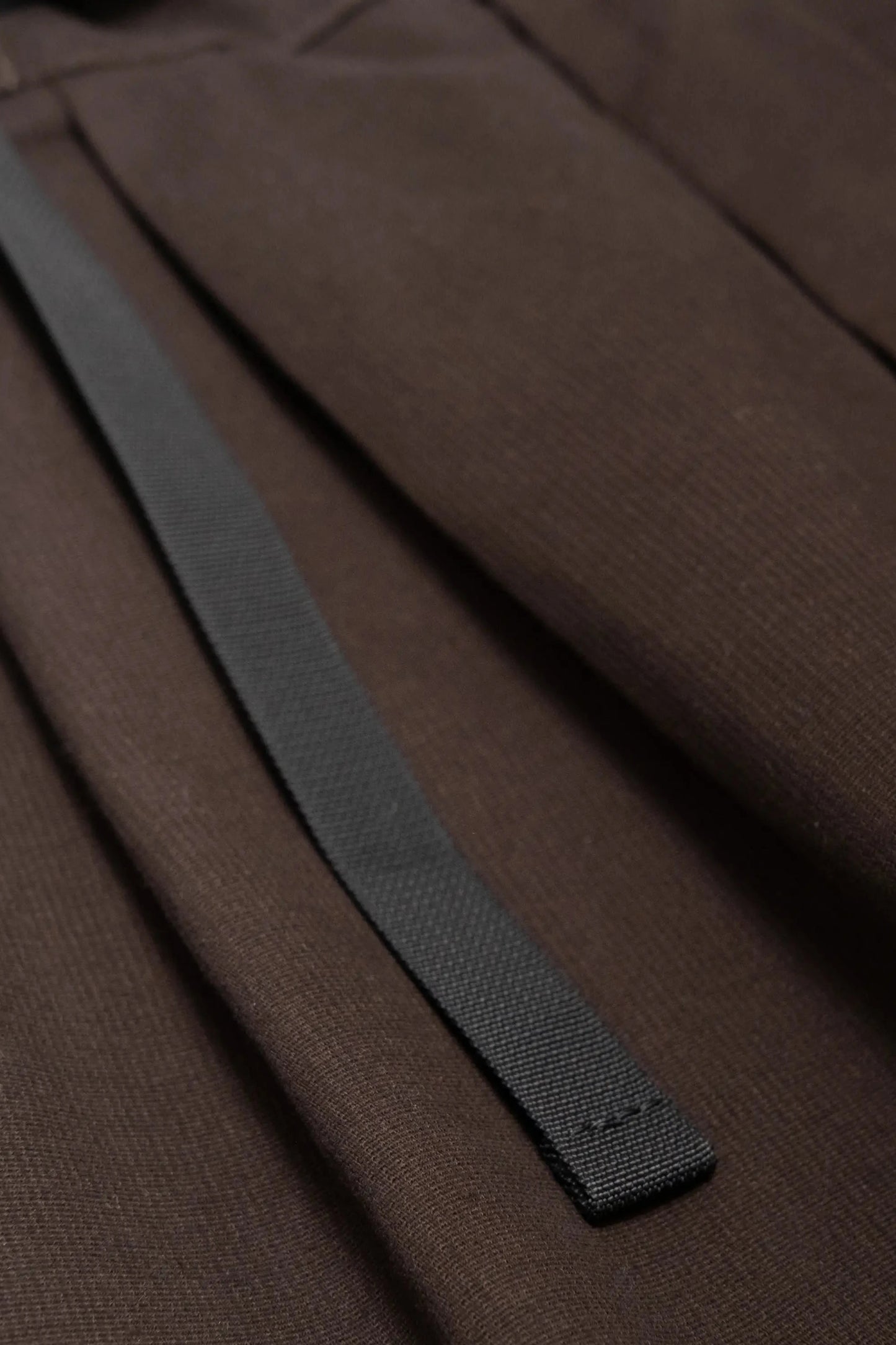 Close-up of brushed brown fabric with a black woven strap detail, highlighting the texture and pleated design for a crafted, utility-inspired look.