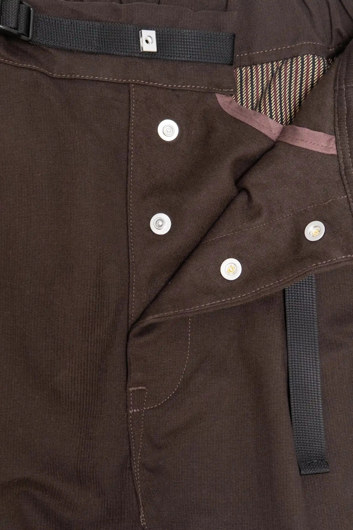 Close-up of dark brown brushed fabric pants with button fly closure, striped inner lining, black adjustable waistband strap, and reinforced stitching details.