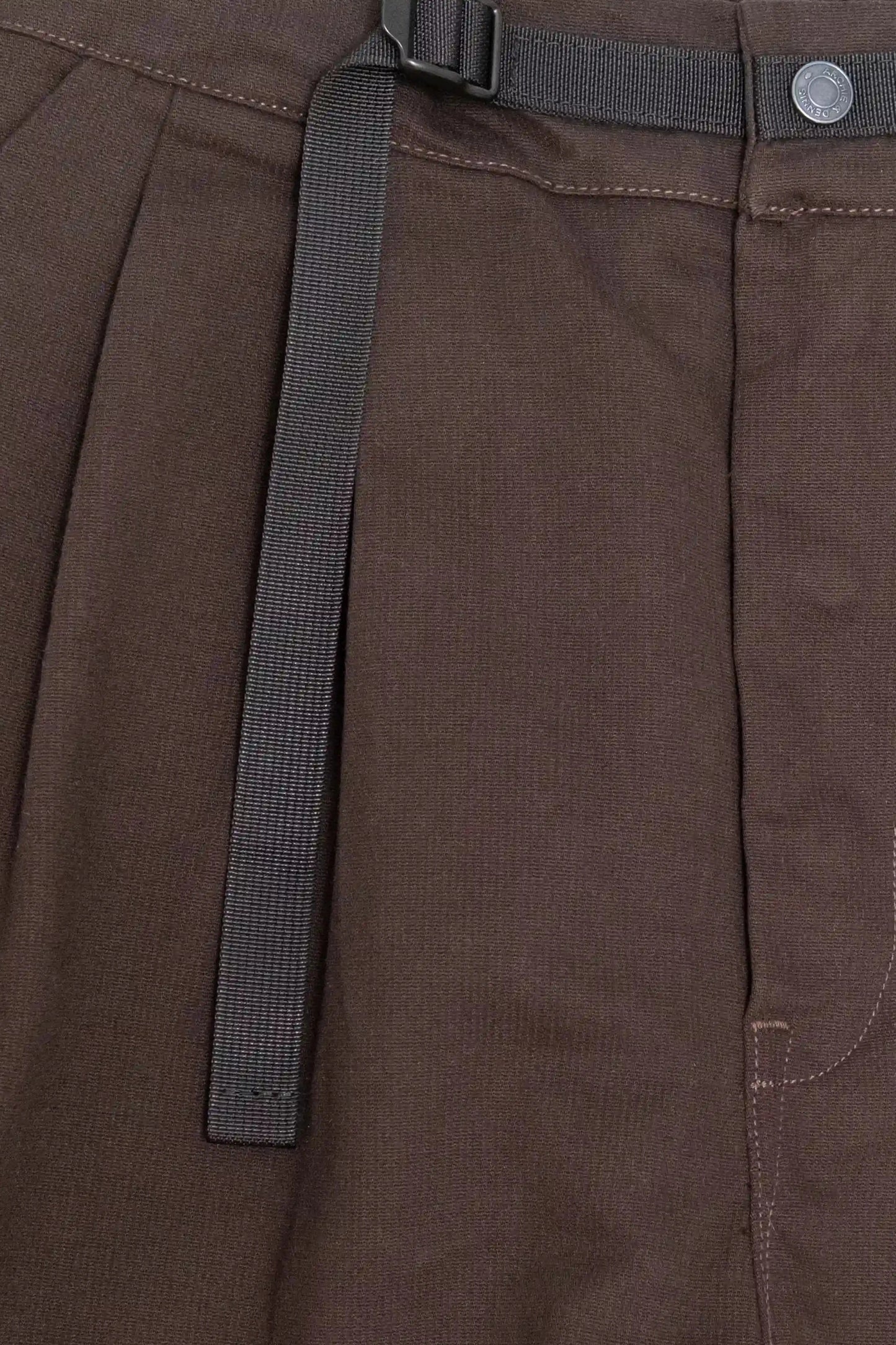 Close-up of brushed brown fabric pants featuring pleated detailing, a black adjustable belt, and a metal button closure with visible stitching for a relaxed, utility-inspired style.