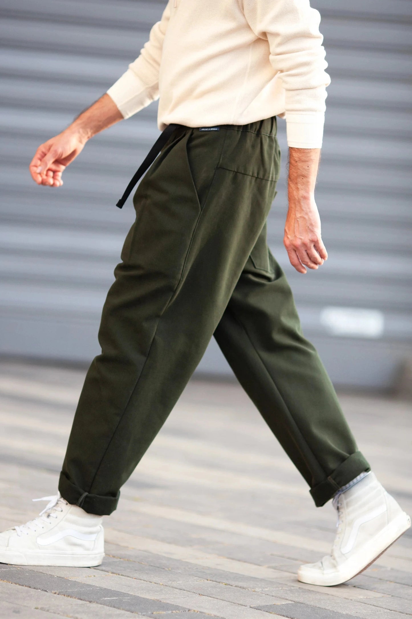 Man wearing relaxed fit dark green pants with a cuffed hem and black belt, paired with white sneakers and a cream sweater, walking outdoors on pavement.