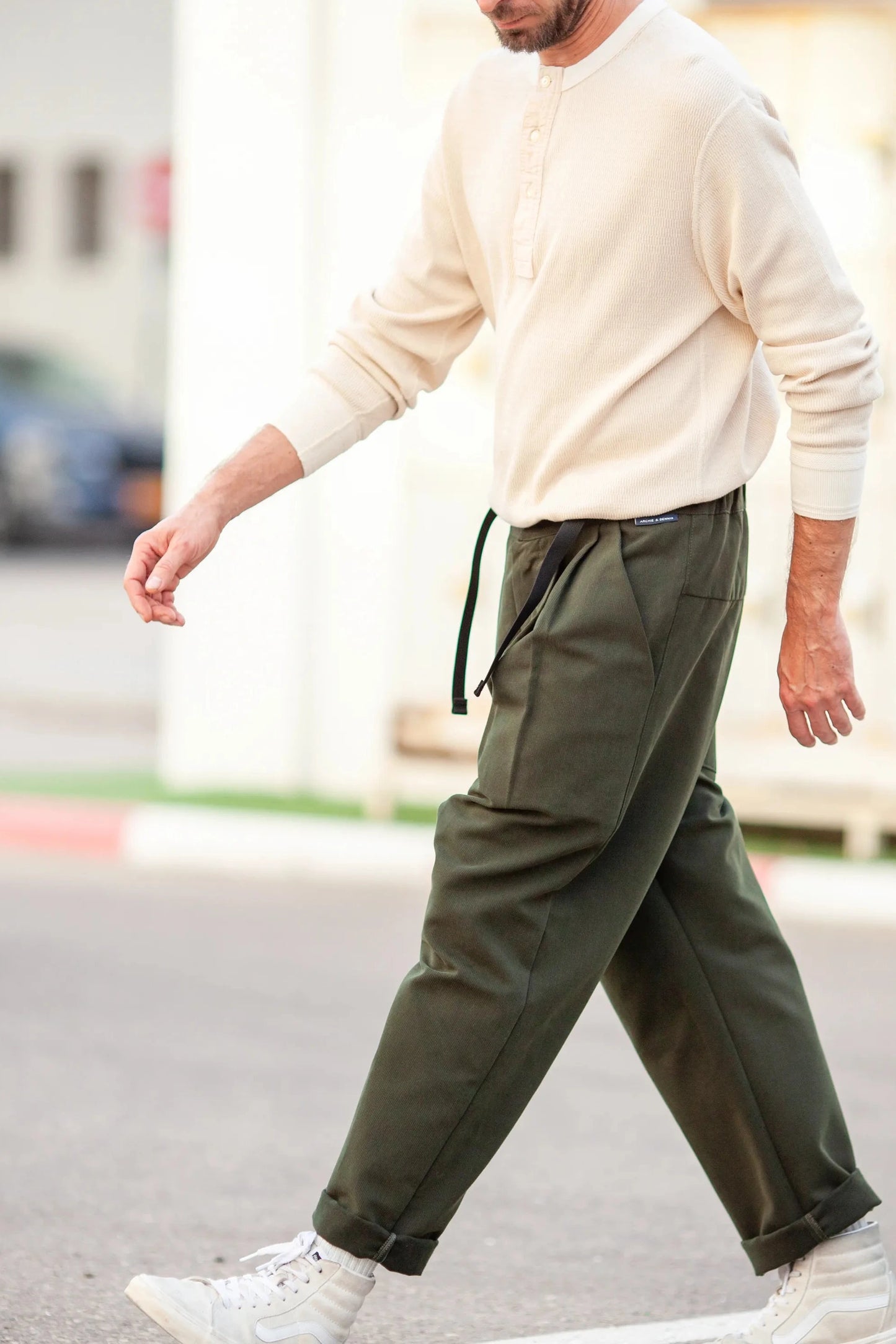Man walking outdoors wearing relaxed fit pleated dark green pants with black drawstring and casual white sneakers paired with a cream long sleeve henley top