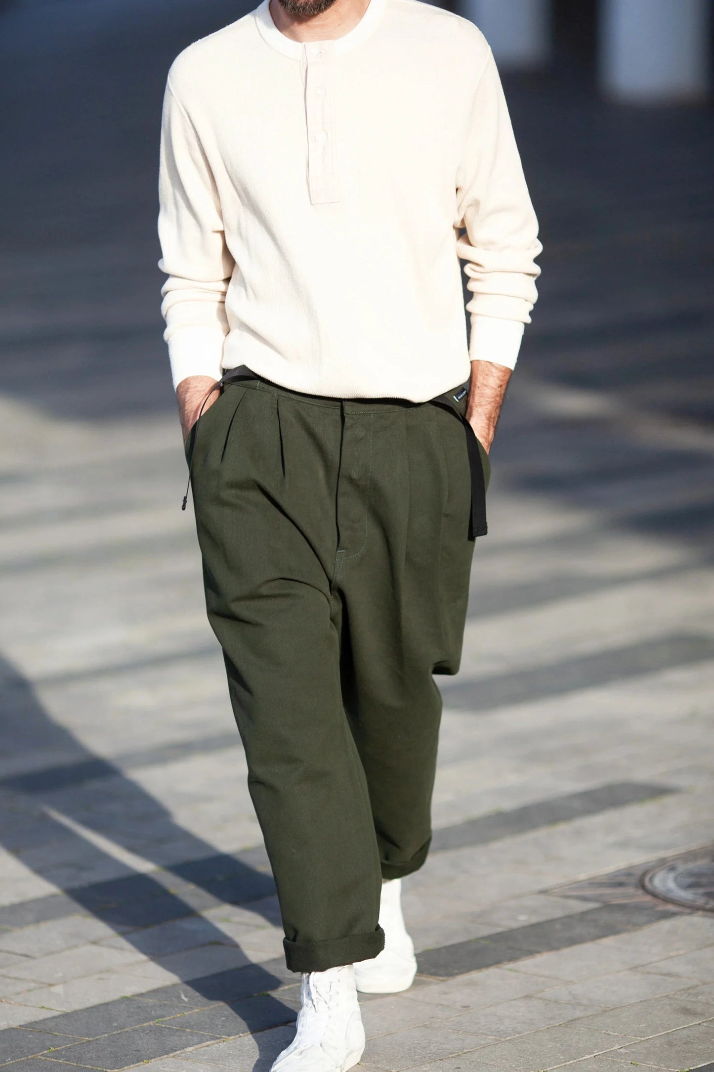 Man wearing relaxed fit, pleated, olive-green pants paired with a casual light cream henley shirt and white sneakers walking outdoors in natural light.