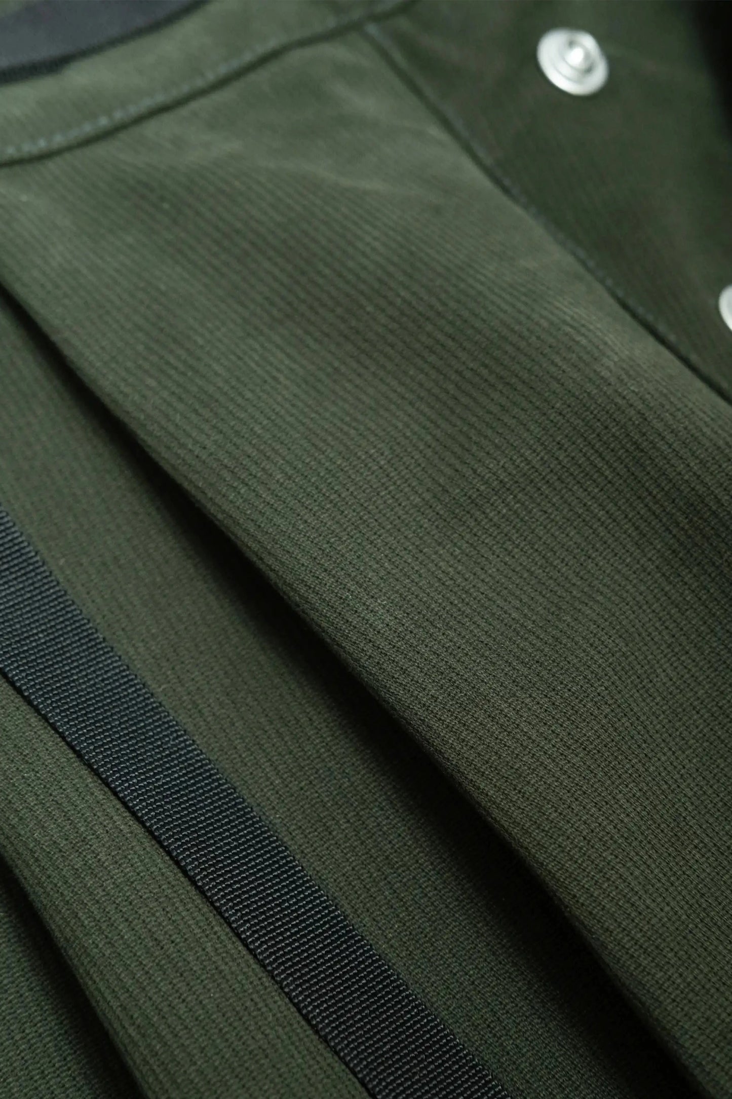 Close-up of textured dark green fabric with pleated design and metallic snap buttons, featuring a black strap detail for utility-inspired menswear style.
