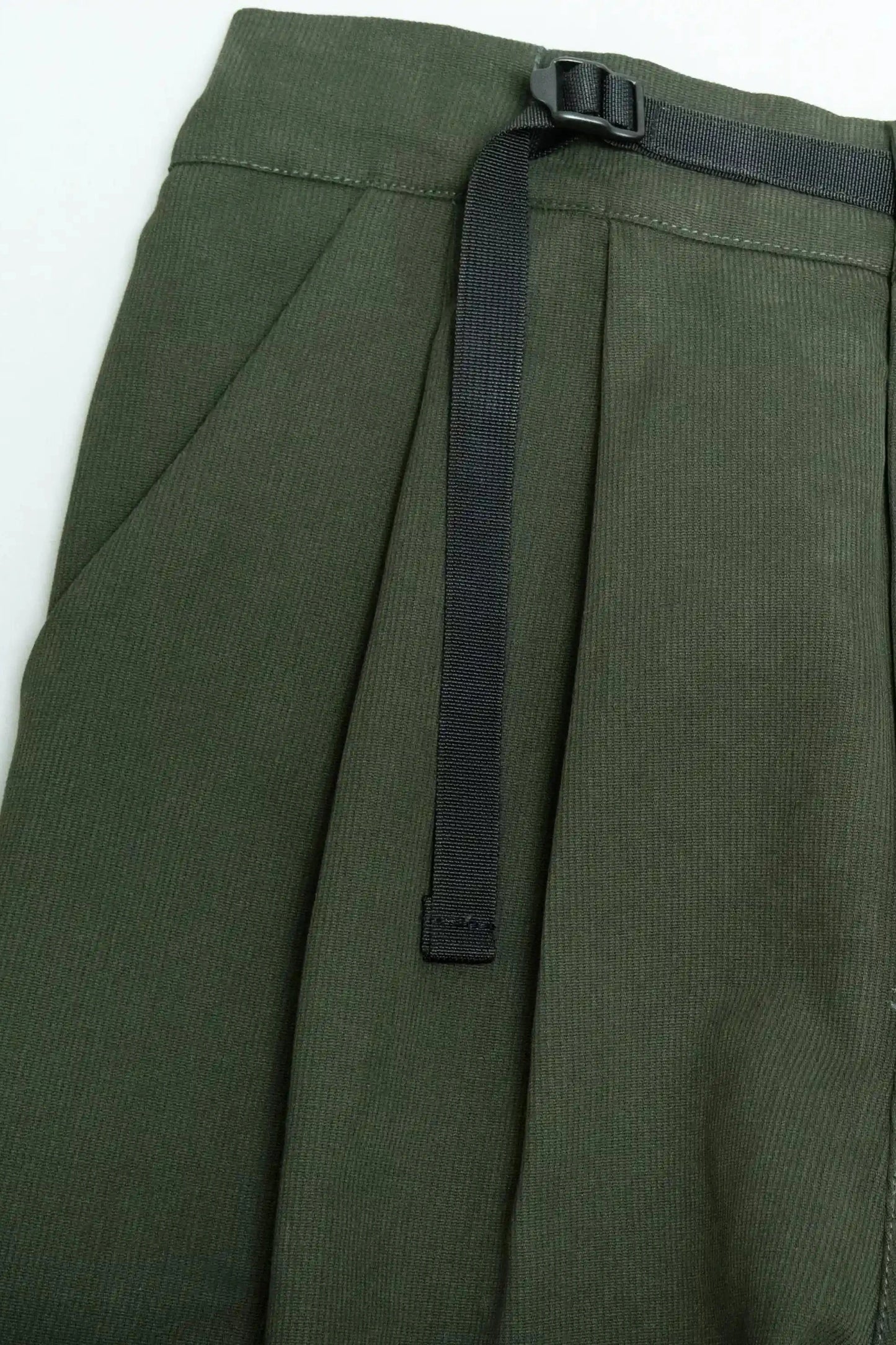 Close-up of olive green pleated pants featuring a black adjustable webbing belt, showcasing a relaxed fit and utilitarian design detail on the waistband.