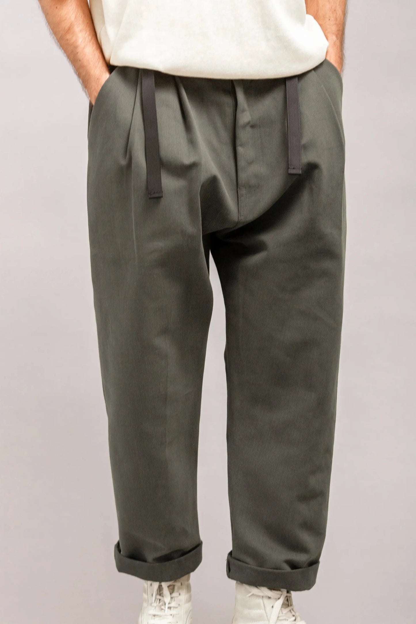 Close-up of relaxed fit olive green pleated pants with black drawstrings and cuffed hems, styled with white sneakers and a tucked-in light beige top.