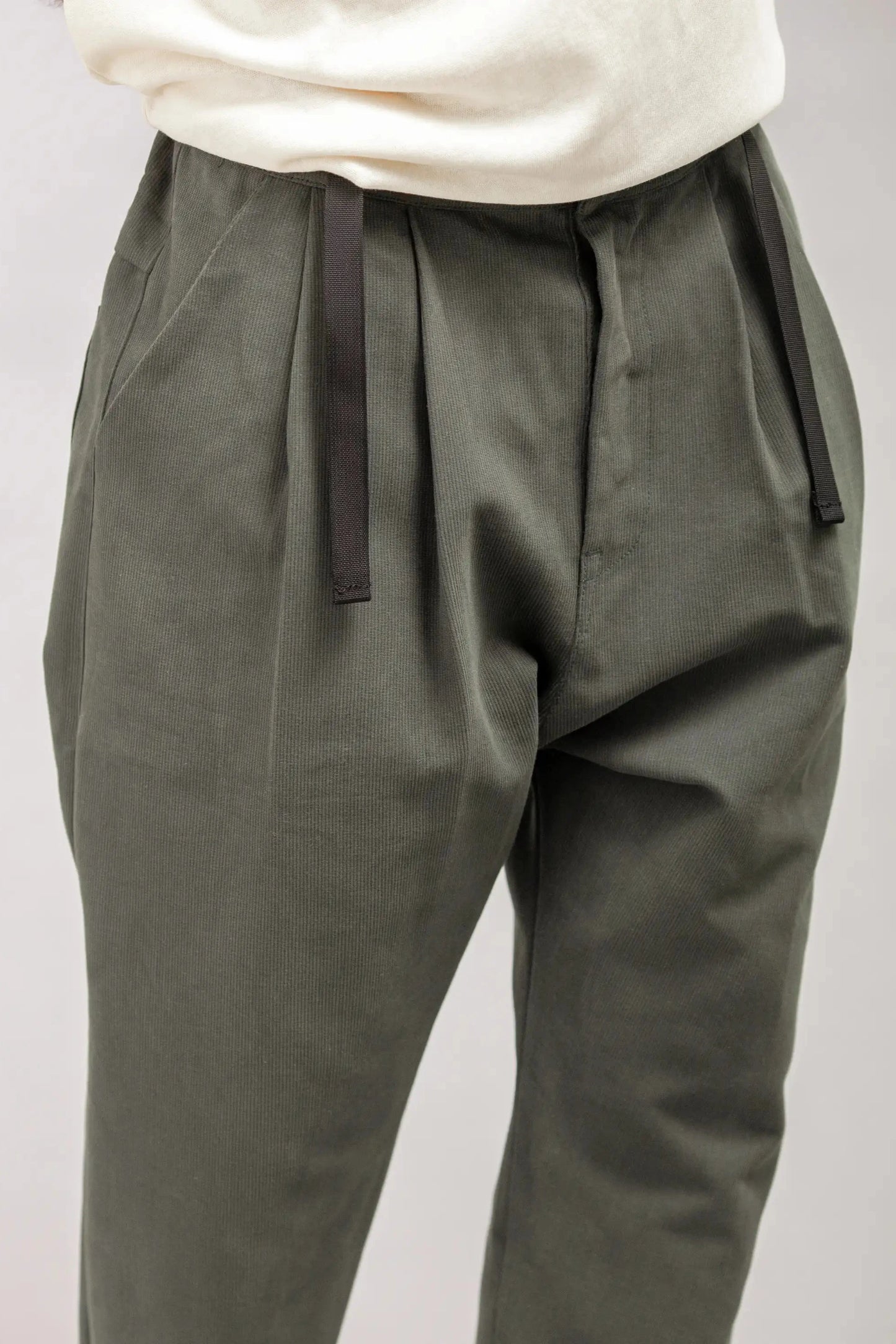 Close-up of relaxed fit pleated pants in muted olive green with black drawstring waistband, styled with casual white top, showcasing utility-inspired menswear design.