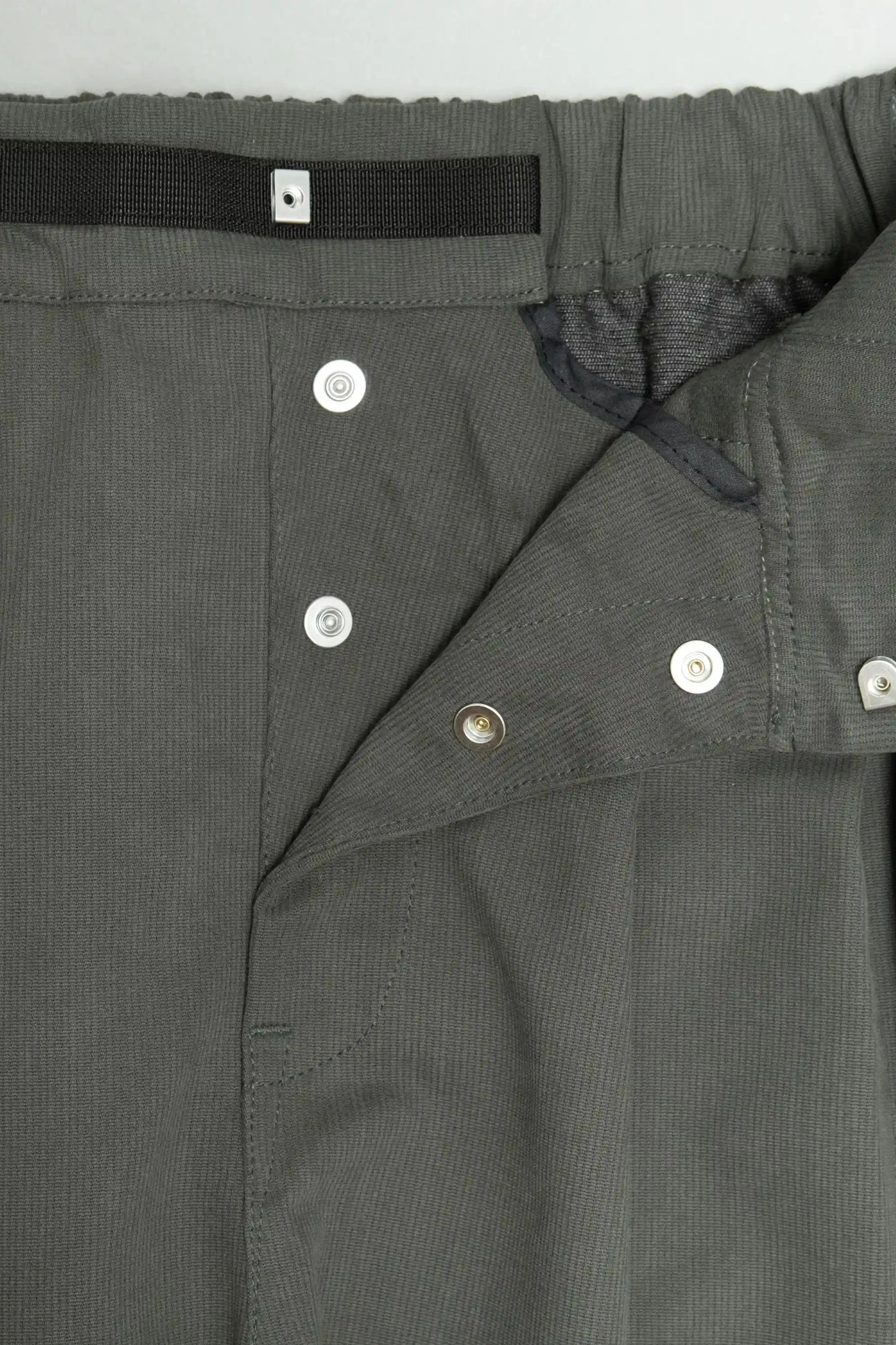 Close-up of dark green utility pants with button closures, elastic waistband, and a black adjustable strap detail, highlighting durable fabric and functional design.