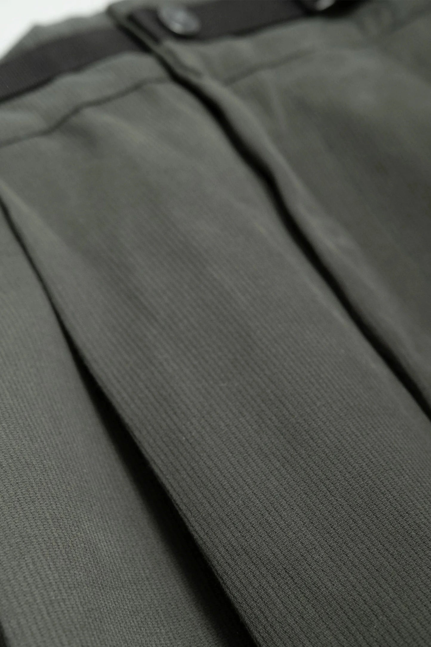 Close-up of olive green pleated fabric detail with black waistband and button, showcasing a military-inspired texture and tailored craftsmanship.