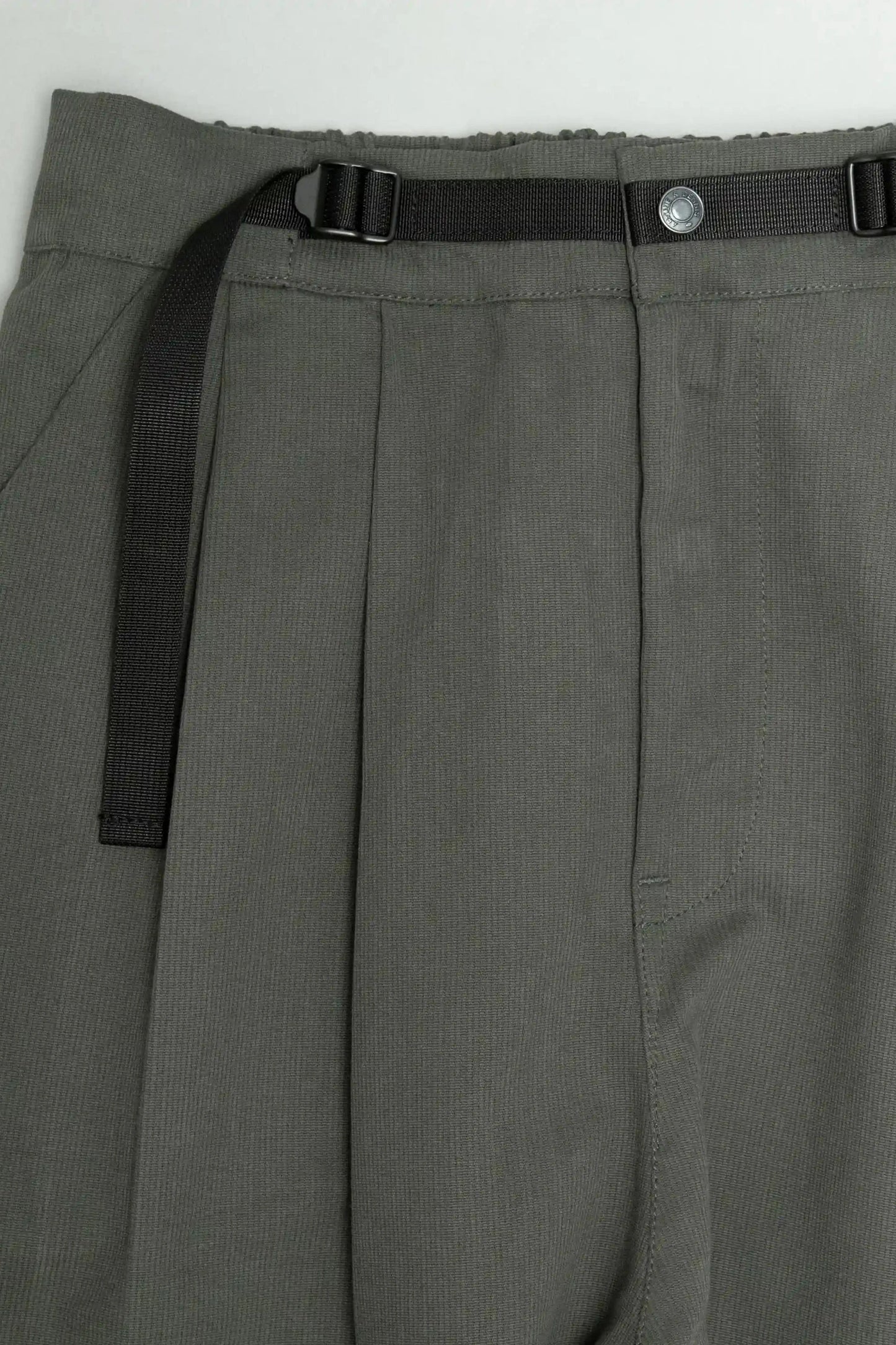 Close-up of olive green pleated pants with a black adjustable webbing belt and button closure, showcasing military-inspired, utility-style design details.