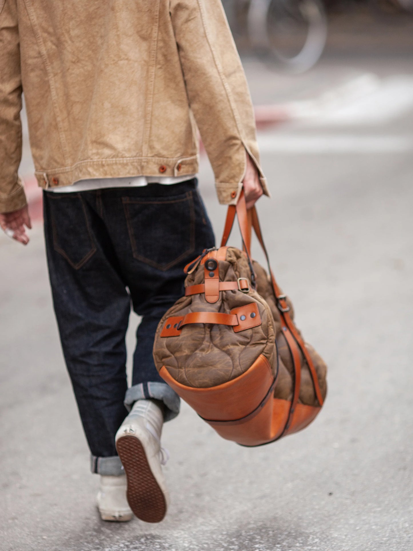 Person walking down a street carrying a brown quilted weekender bag with leather straps, wearing dark rolled-up jeans, white shoes, and a light tan jacket.