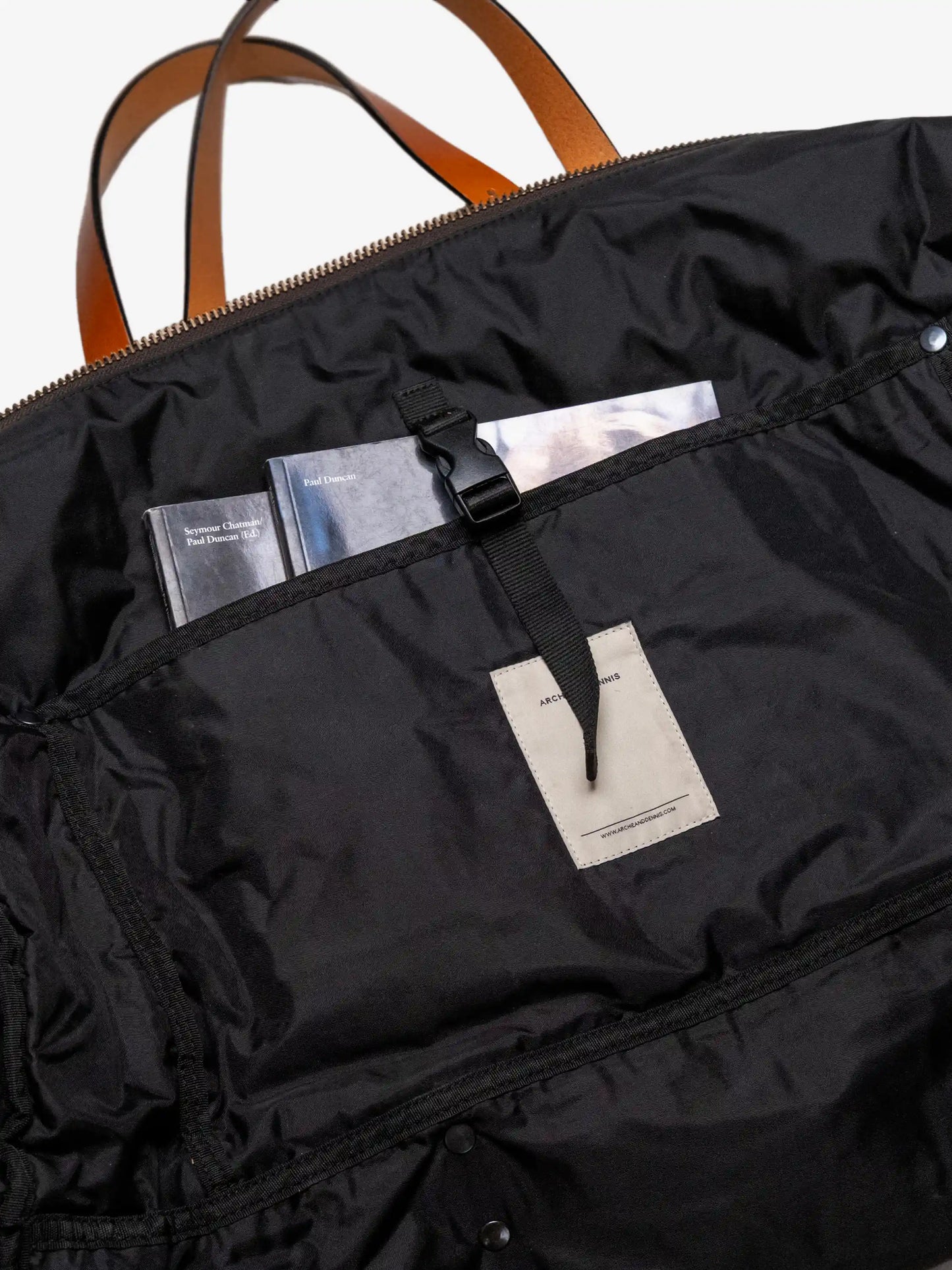 Interior view of a black quilted weekend bag with leather handles, showing an organized inner pocket secured with a black strap holding two books.