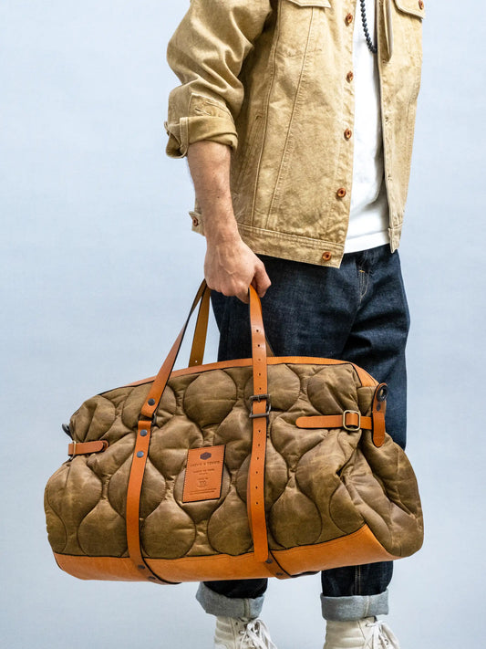 Man holding a quilted weekend bag with leather straps, styled with casual denim jeans and a beige jacket, highlighting a relaxed and utility-inspired look.
