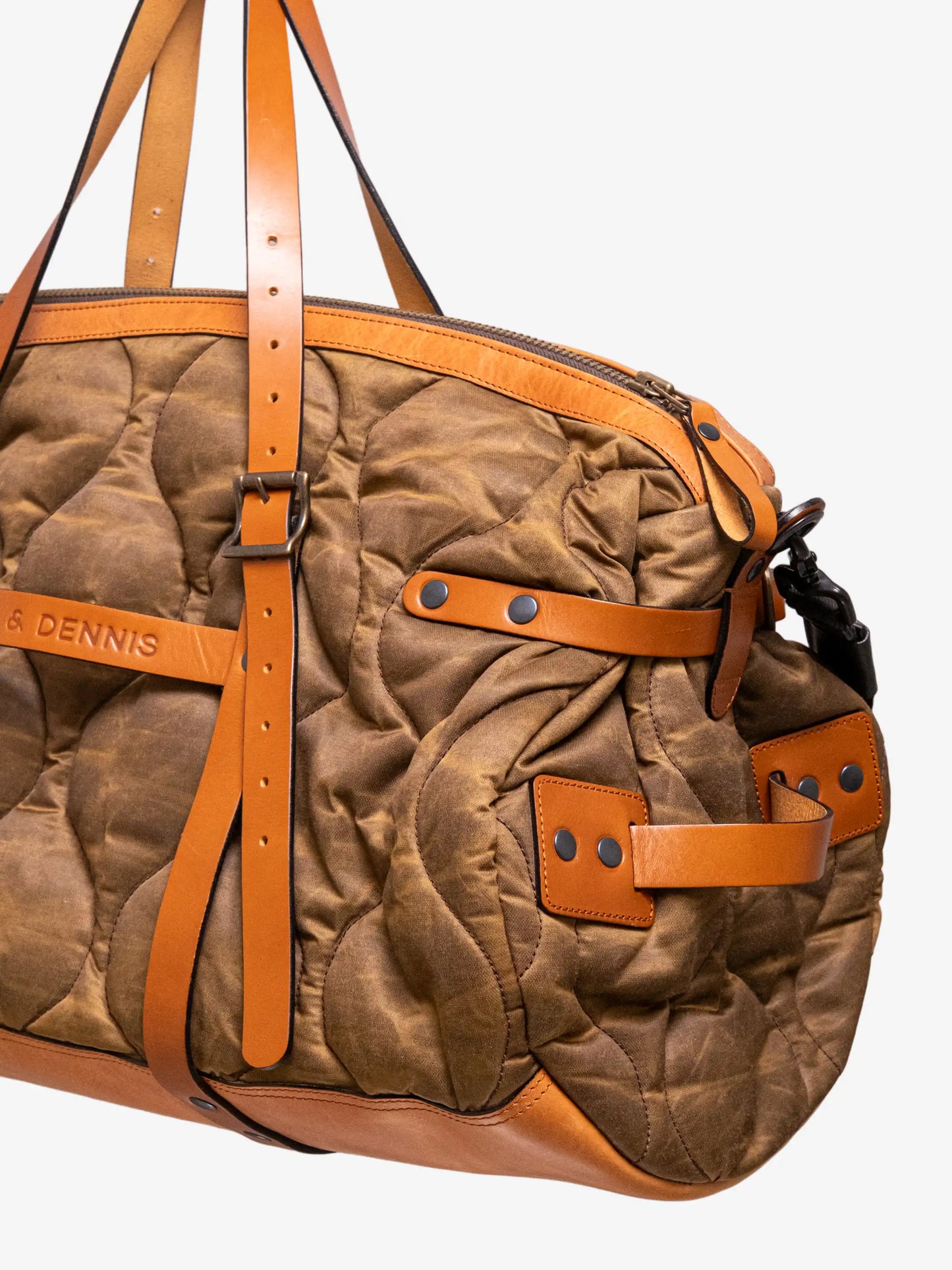 Close-up of a quilted canvas weekender bag with tan leather straps, brass hardware, and reinforced corners designed for stylish, durable travel.