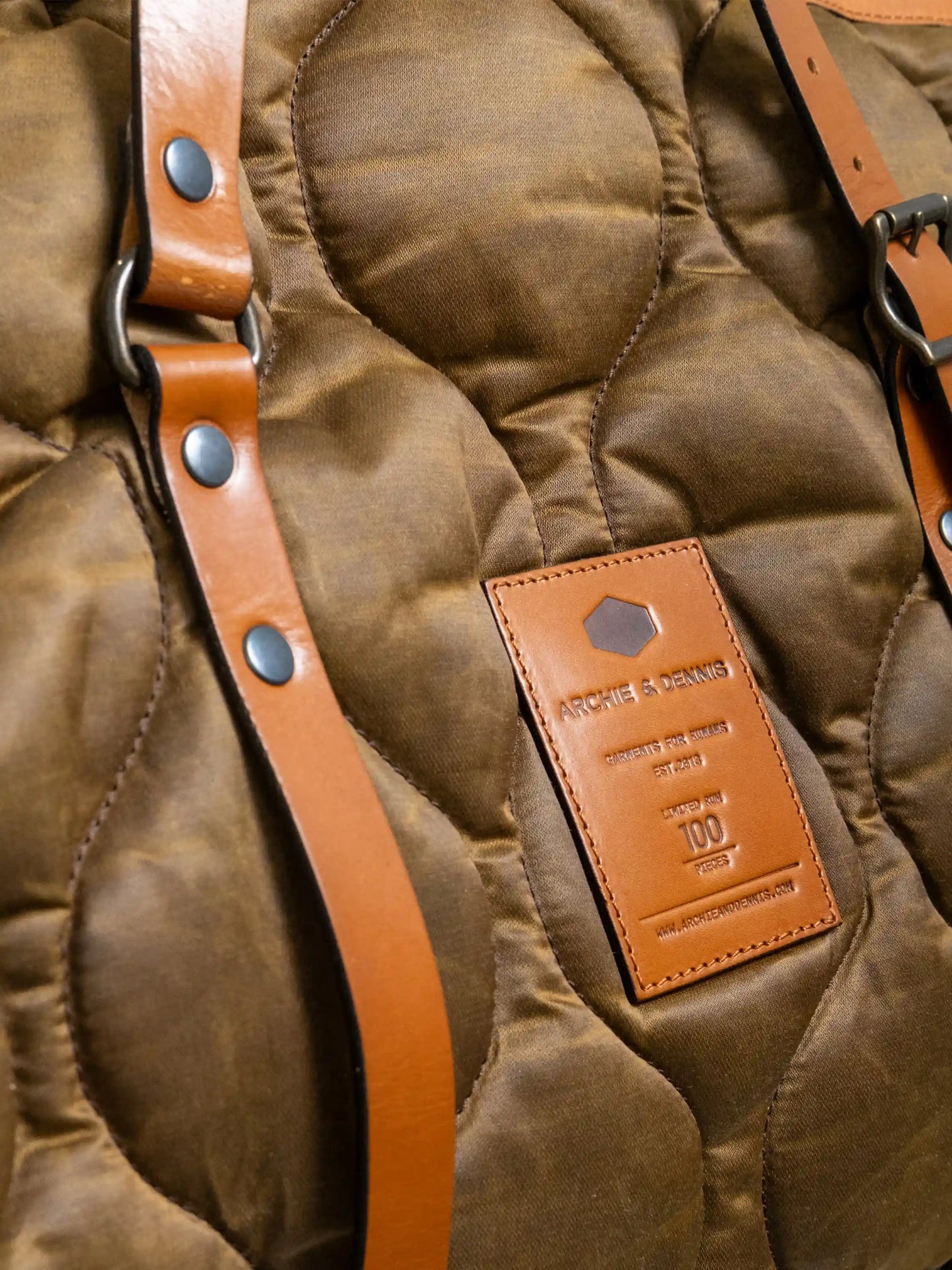 Close-up of a quilted fabric bag with tan leather straps and a stitched leather label featuring branding and design details in embossed text.