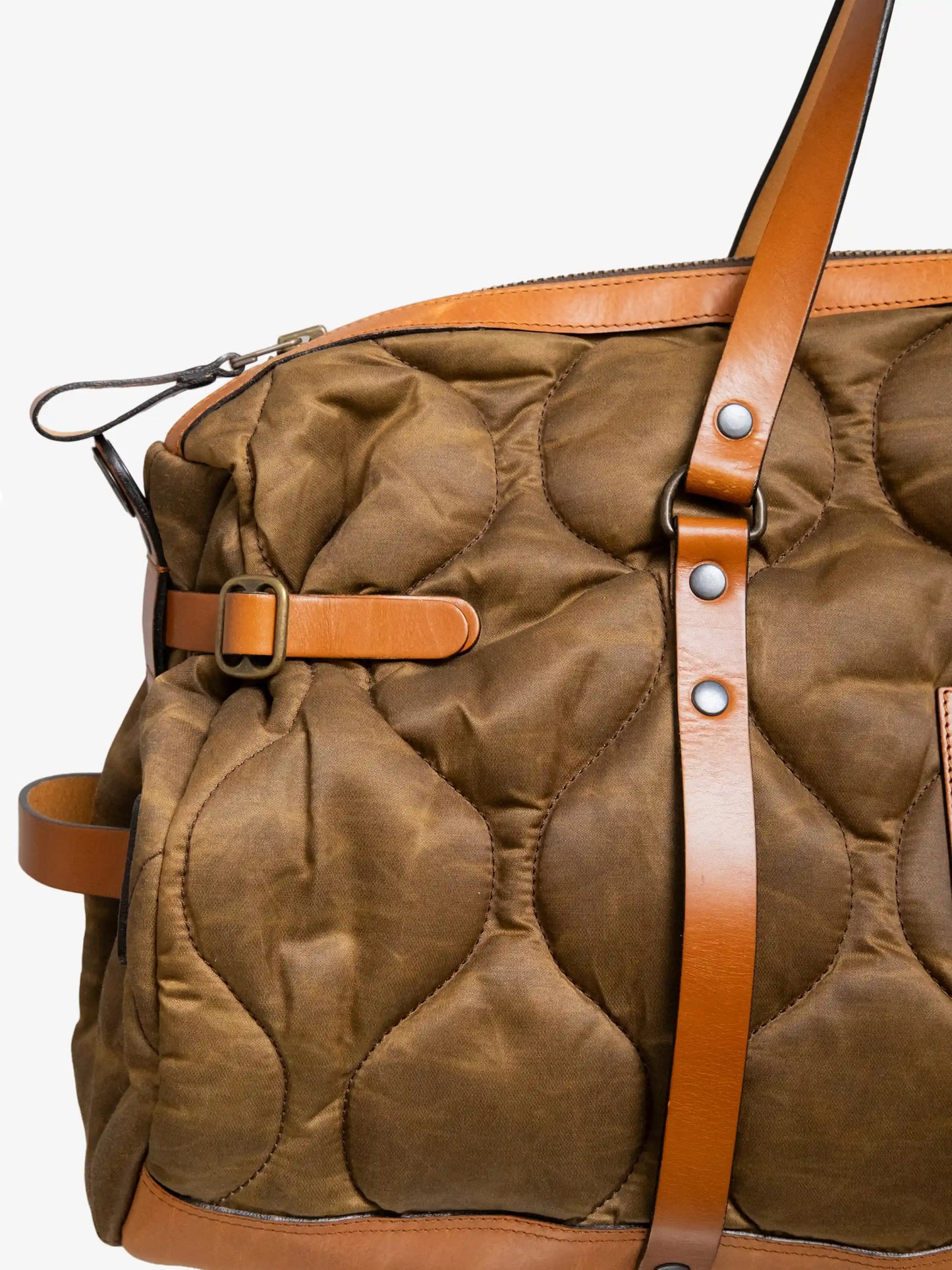 Close-up of a quilted weekend bag in brown fabric with tan leather straps and metal buckle details, showcasing craftsmanship and utility-inspired design.
