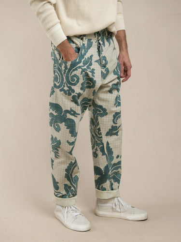 Man wearing relaxed fit pants with a blue-green floral and scroll pattern on a light background, paired with white sneakers and a cream-colored sweater.