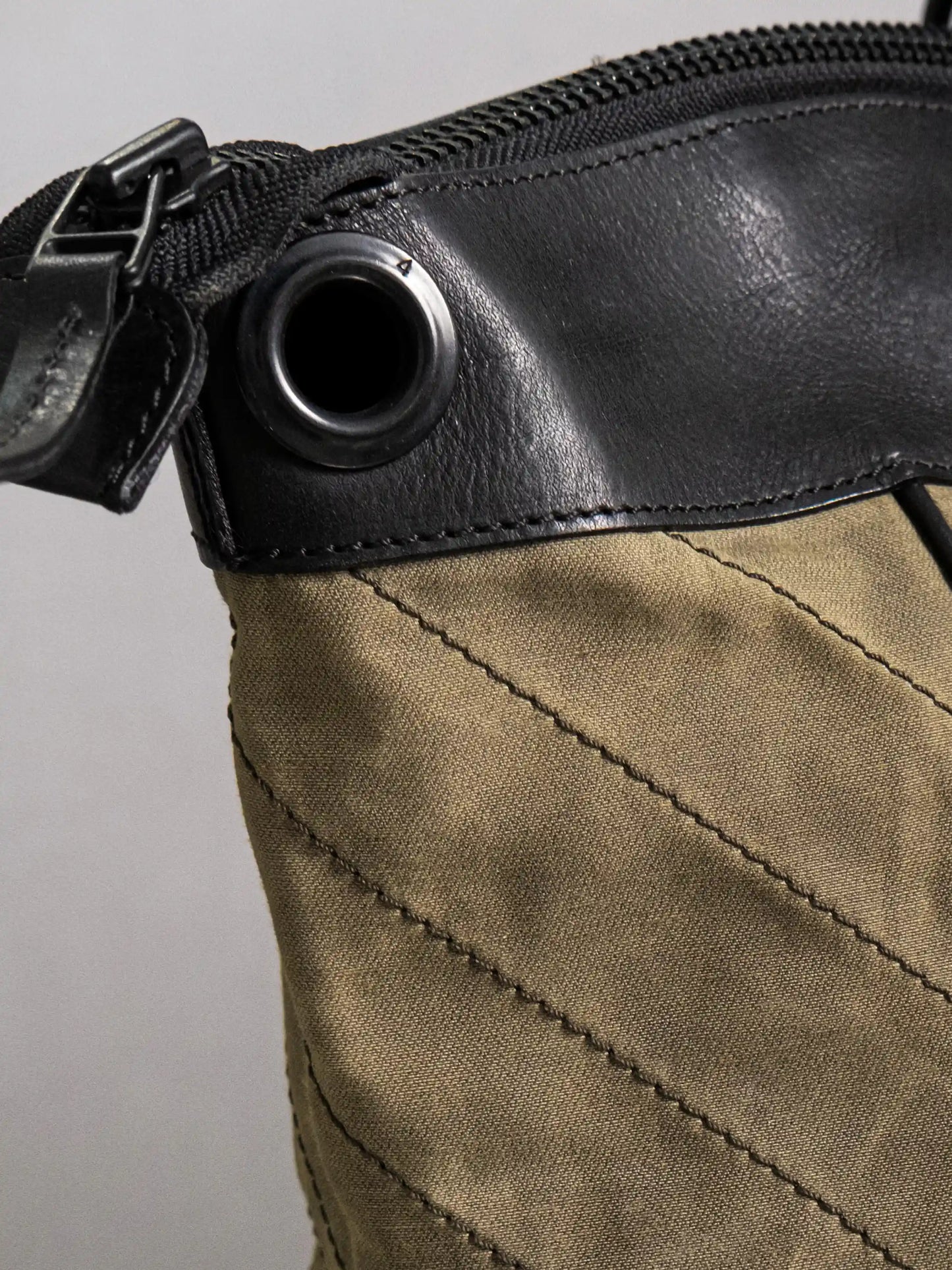 Close-up of a handmade olive canvas bag featuring black leather trim, metal grommet, and detailed stitching along the fabric seams.