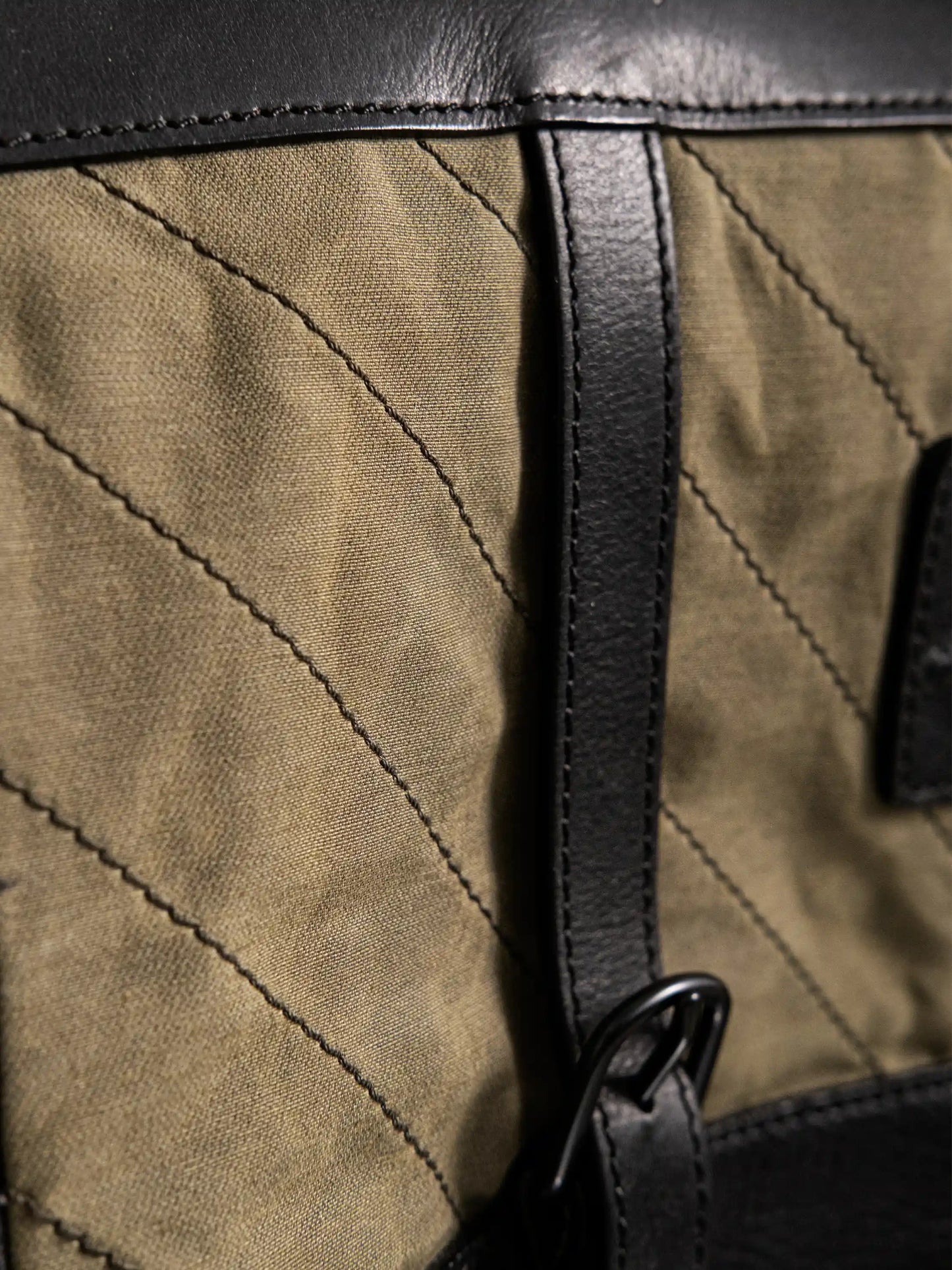 Close-up of an olive fabric bag with black leather trim and strap detailing, featuring diagonal stitching for a handcrafted, military-inspired look.